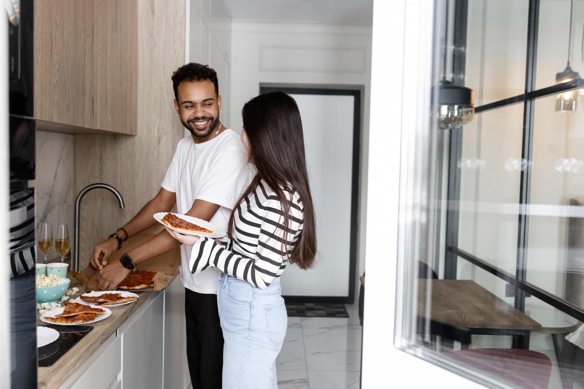 A man and woman stand in a kitchen, surrounded by various food items on the counter and stove.
